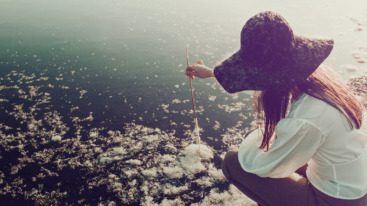Woman in a big hat draw on the water with a stick and reeds feathers