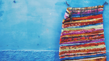 Een geweven wandkleed in verschillende kleuren tegen een blauwe achtergrond.