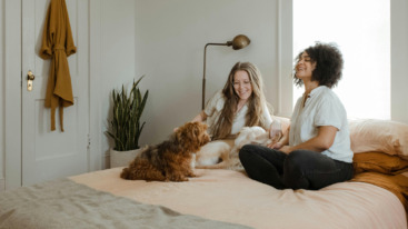 twee vrouwen die samen met een hond op bed zitten en lachen.