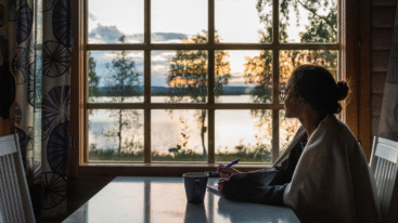 Een dame met een pen in haar hand en een notititeblok voor zich, starend uit het raam met een mooi uitzicht op een meer.